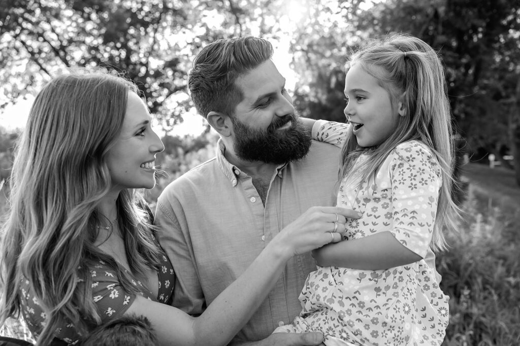 mom tickling little girl while dad holds her and smiles at her during family photos outside